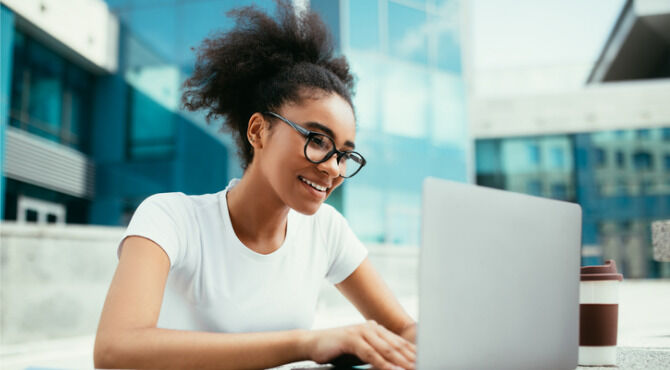 African American College Girl Using Laptop Learning Online And Browsing Internet Doing Homework In Modern University Outdoors. Student Typing On Computer, Studying Wearing Eyeglasses. Side View