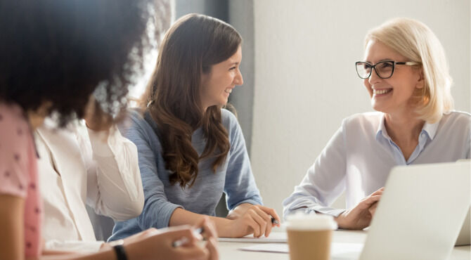 Friendly smiling old and young businesswomen talking laughing at corporate group business meeting, happy female employees colleagues coach manager and intern having fun pleasant conversation at work