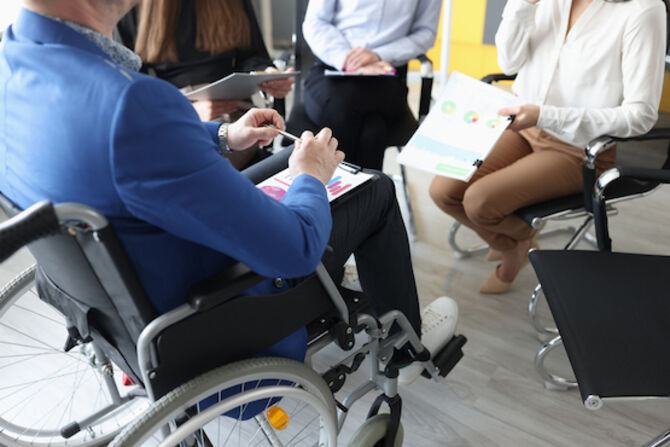 Disabled man with documents in hands sitting in wheelchair against background of colleagues closeup. Adaptation of disabled people and success to work concept