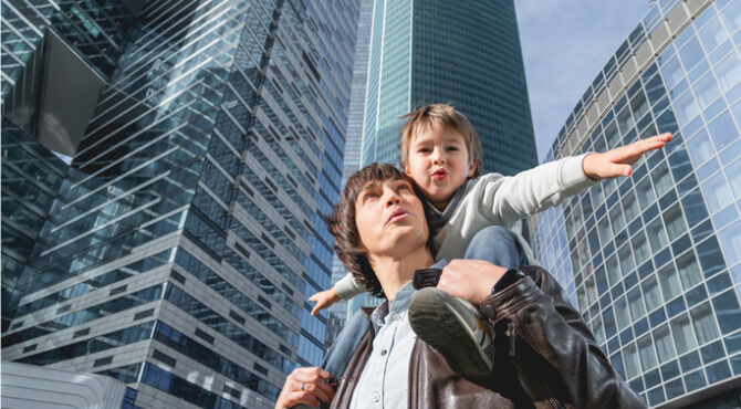 Laughing boy plays like airplane on father's shoulders. Dad and son looks on glass walls of buildings. Future and modern technologies, life balance and family life in well keeps districts.