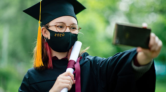 A young woman graduate, wearing a mortar board and holding her diploma, takes a selfie