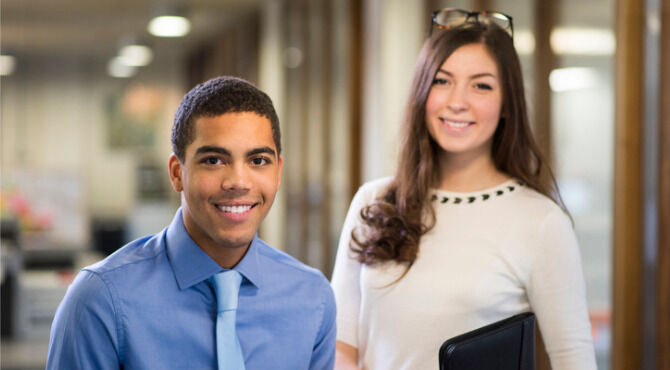 Young man and woman stand in their new workplace smiling proudly to camera