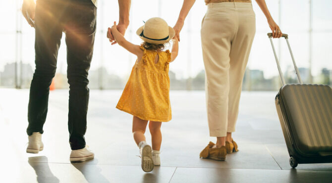 Back view of husband and his wife holding hands and walking with their little girl and suitcase at the airport.