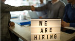 Close up of help wanted sign during a job interview in the office with people in the background.