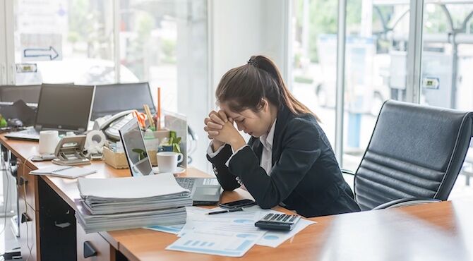 Woman at desk with head in her hands, illustrating an article about welllness