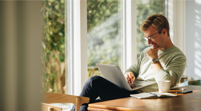 Young man using laptop and smiling at home. Man sitting by table working on laptop computer.